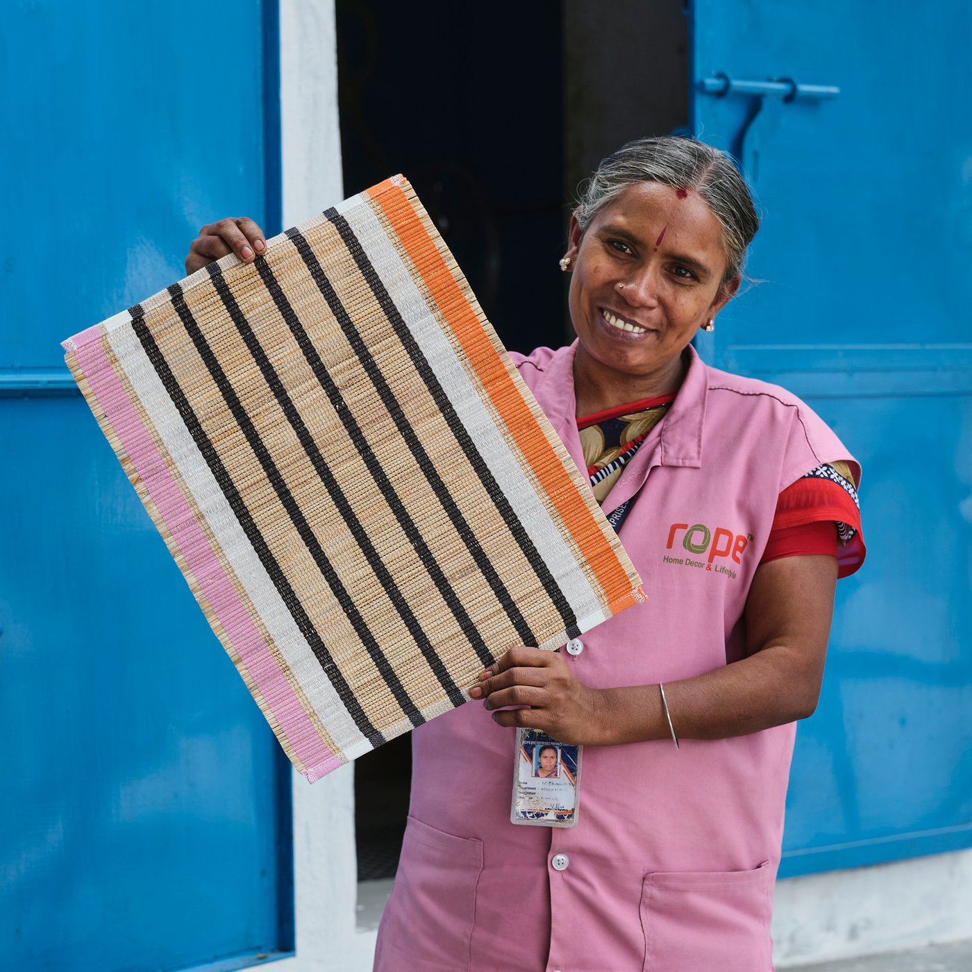A person in pink clothes stands smiling in front of a pair of blue doors and holds up a striped MÄVINN place mat.