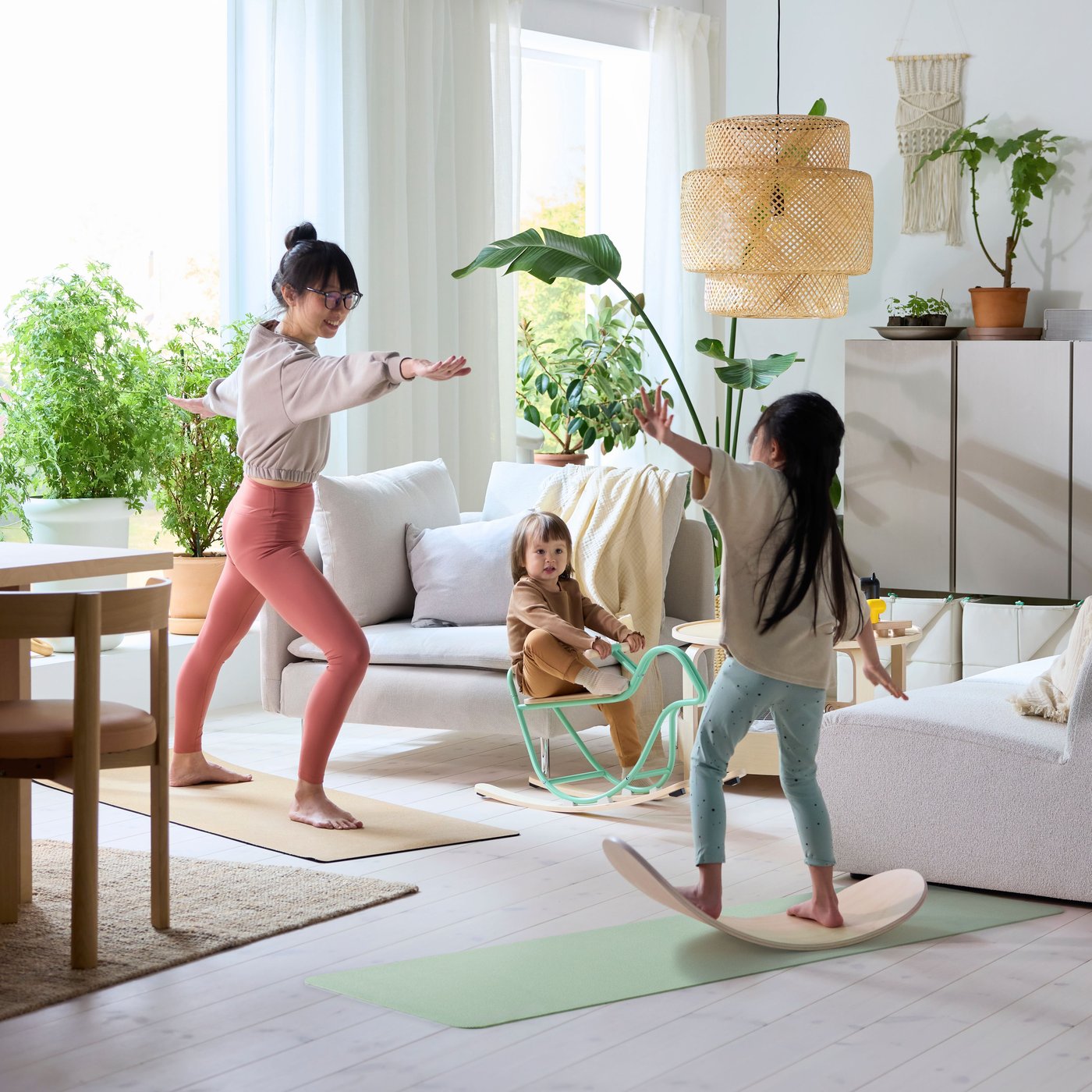 I ett vardagsrum utövar en förälder yoga medan barnen leker på en GREJSIMOJS balansbräda och en GREJSIMOJS gunghästfågel.