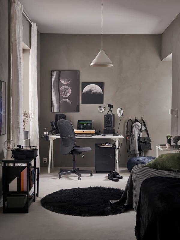 A grey bedroom with a home studio work desk and chair, laptop and speakers, OBEGRÄNSAD black rug, bed and shelving unit.