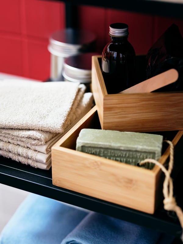 Des serviettes pliées, des bocaux et des bouteilles, et des accessoires de salle de bain dans deux plateaux TAVELÅN sont placés sur l’étagère d’un rangement ENHET.