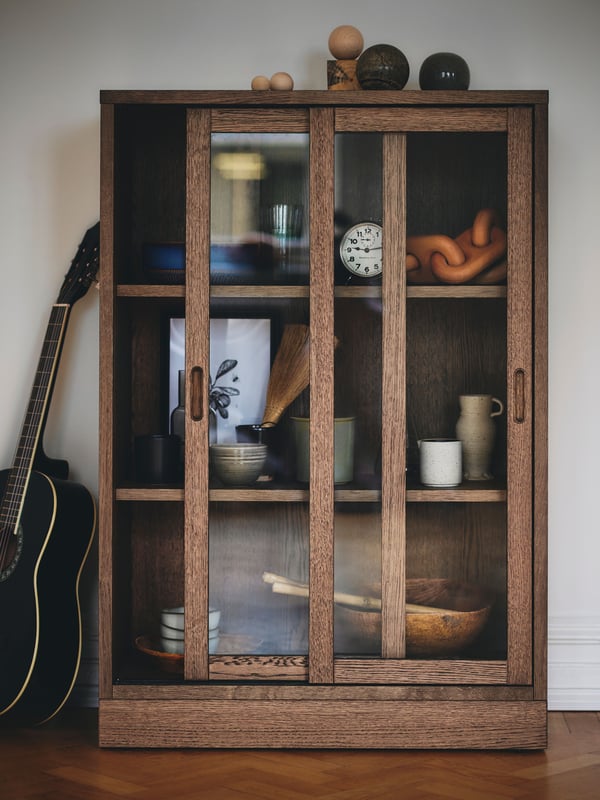 A brown stained oak veneer TONSTAD cabinet with sliding glass doors stands against a white wall with a guitar next to it.