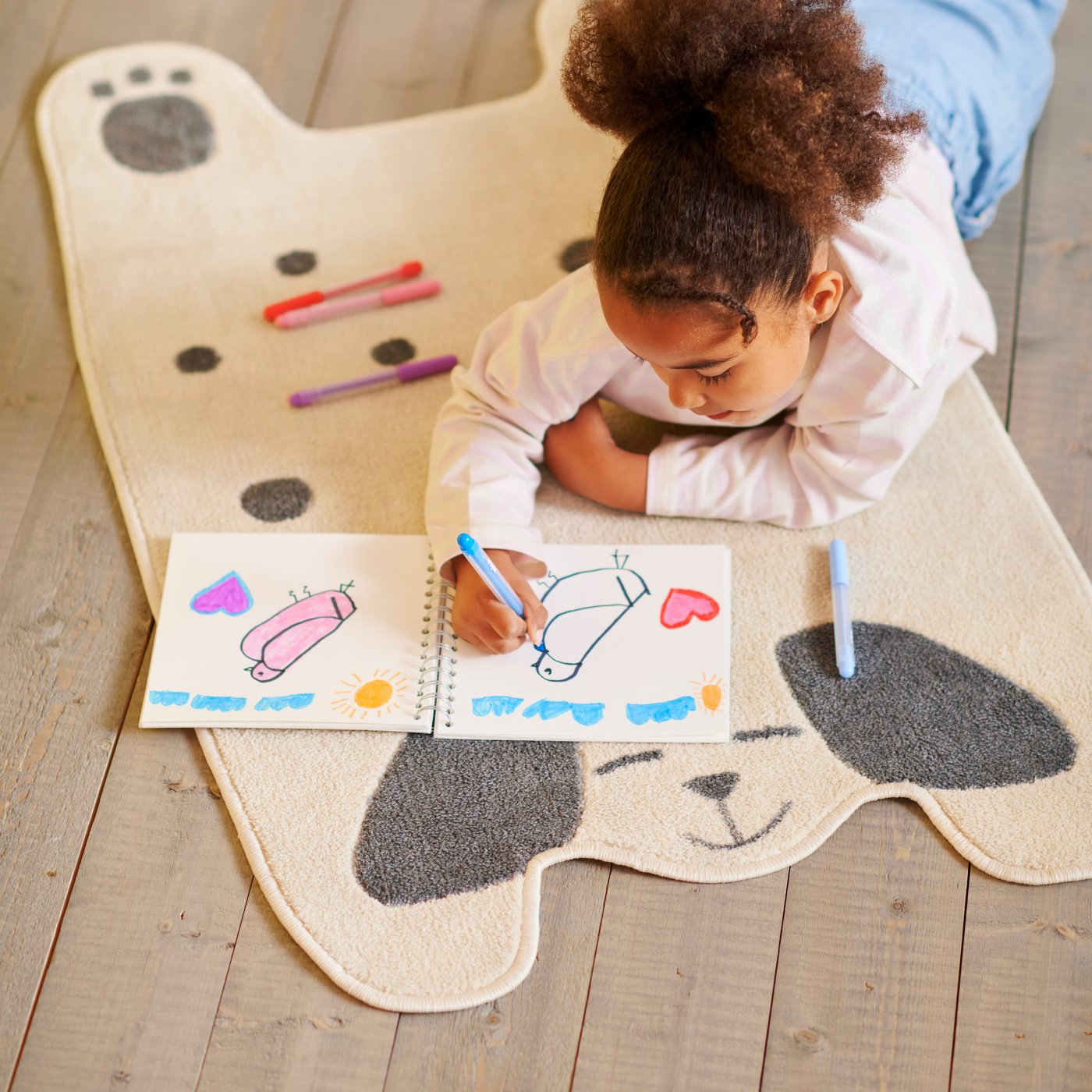 A child is drawing in a notebook using colourful pens and smiling, lying on a DRÖMSLOTT dog-shaped rug.