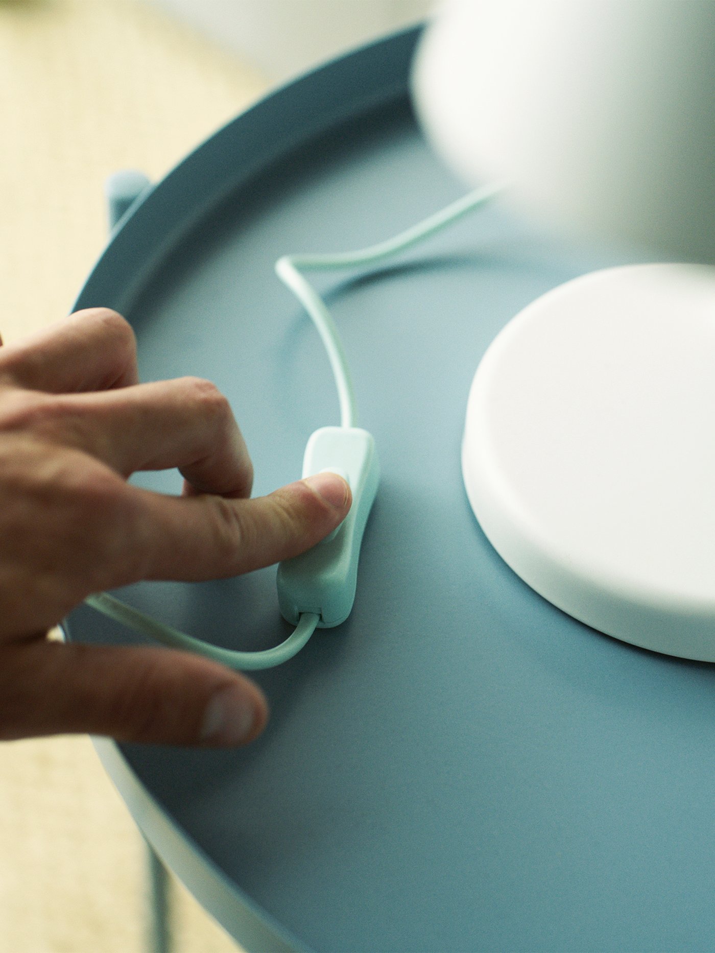 A hand pressing the switch of a white table lamp standing on a light blue table.