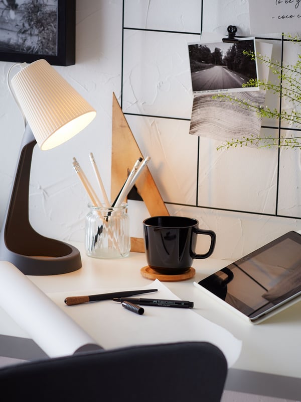 Photos and quotes on a black SÖSDALA memo board with clips above a desk with a SVALLET work lamp, a mug, and a tablet stand.