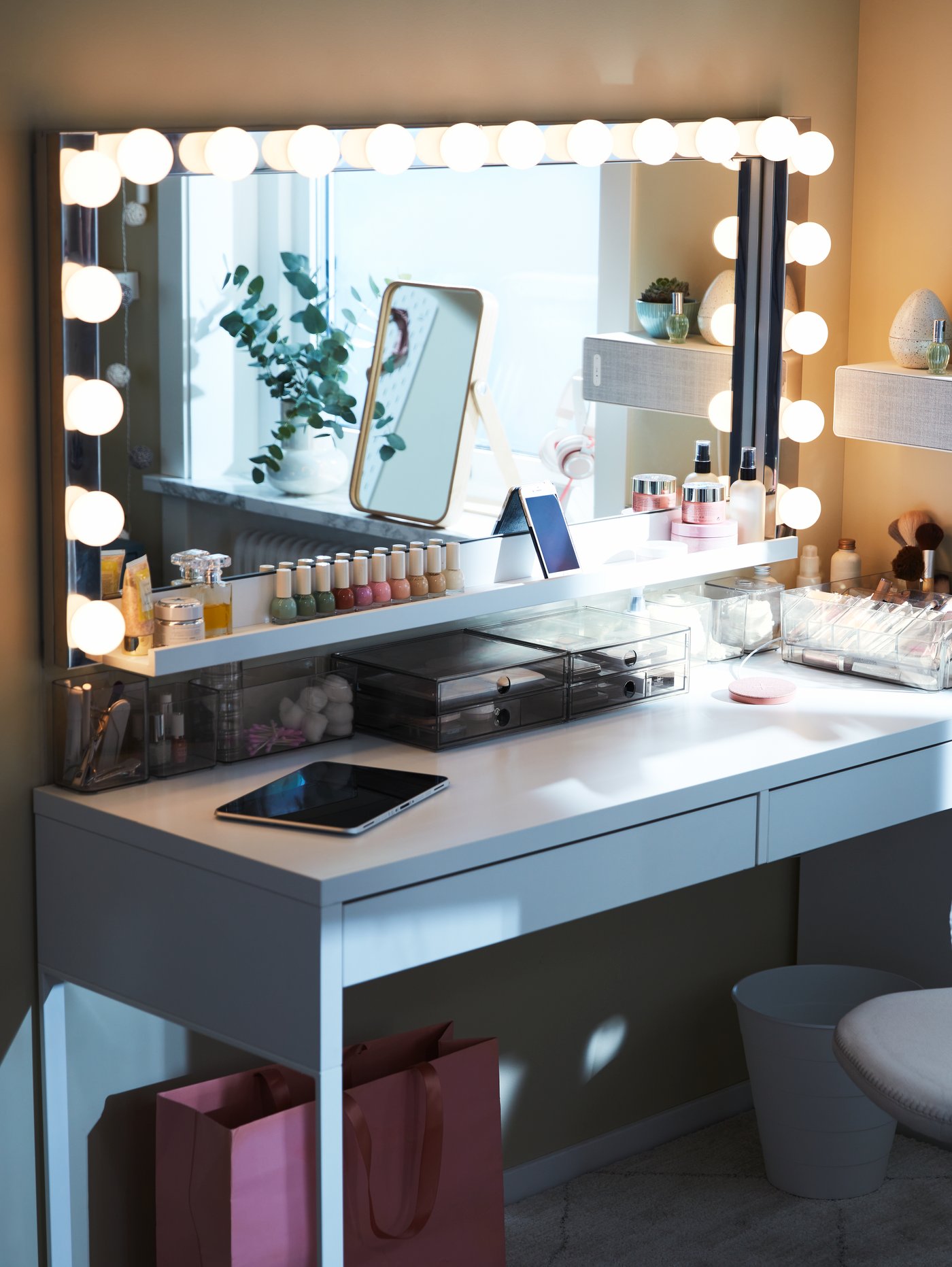 Dressing table with a large mirror surrounded by light bulbs, makeup products and a tablet placed on the table. Makeup storage inspiration.