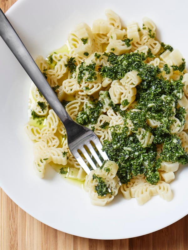 A fork in stainless steel and white bowl containing BÄSTISAR organic pasta, parsley and olive oil, on a wooden surface.