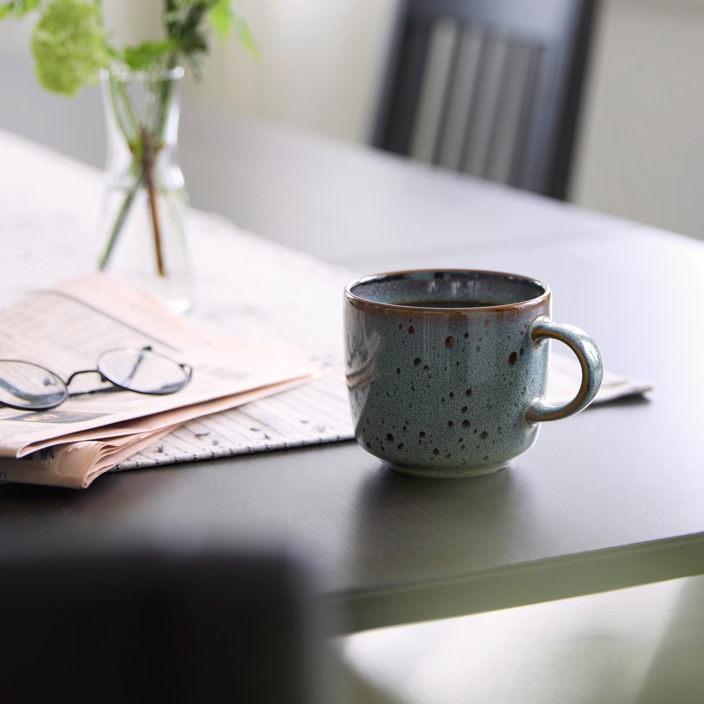 Mug GLADELIG beige/vert posé sur une table près d’un journal et d’une paire de lunettes. Vase garni de verdure en arrière-plan.
