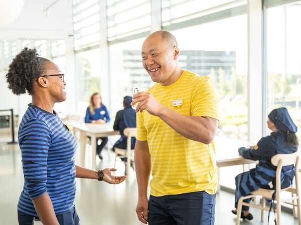 Two IKEA co-workers, one in a blue striped shirt and one in a yellow striped shirt, chat in a bright IKEA staff restaurant.