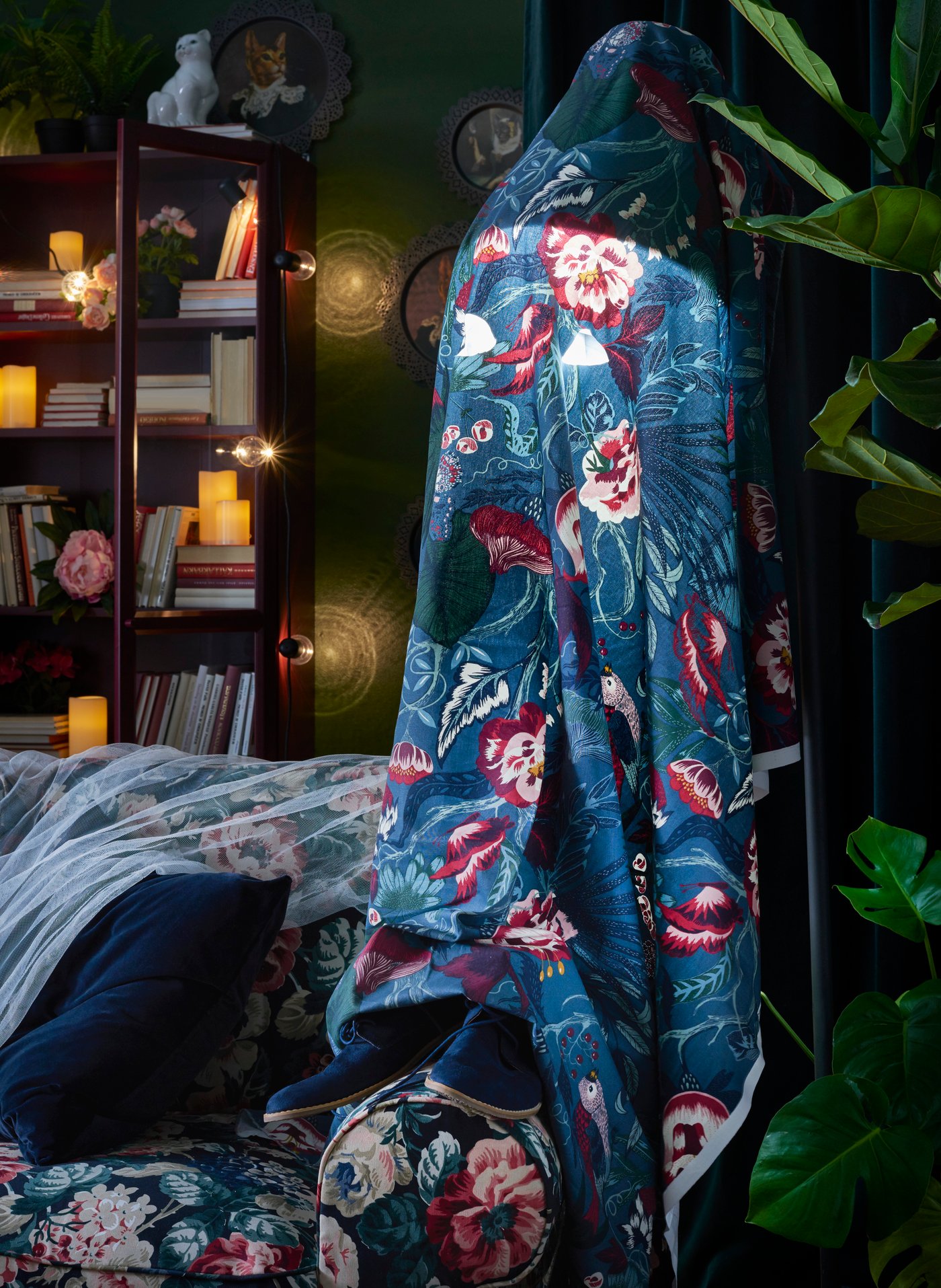 A large floor lamp draped in a dark blue and red floral fabric in a darkly decorated living room with a red bookcase in the background.