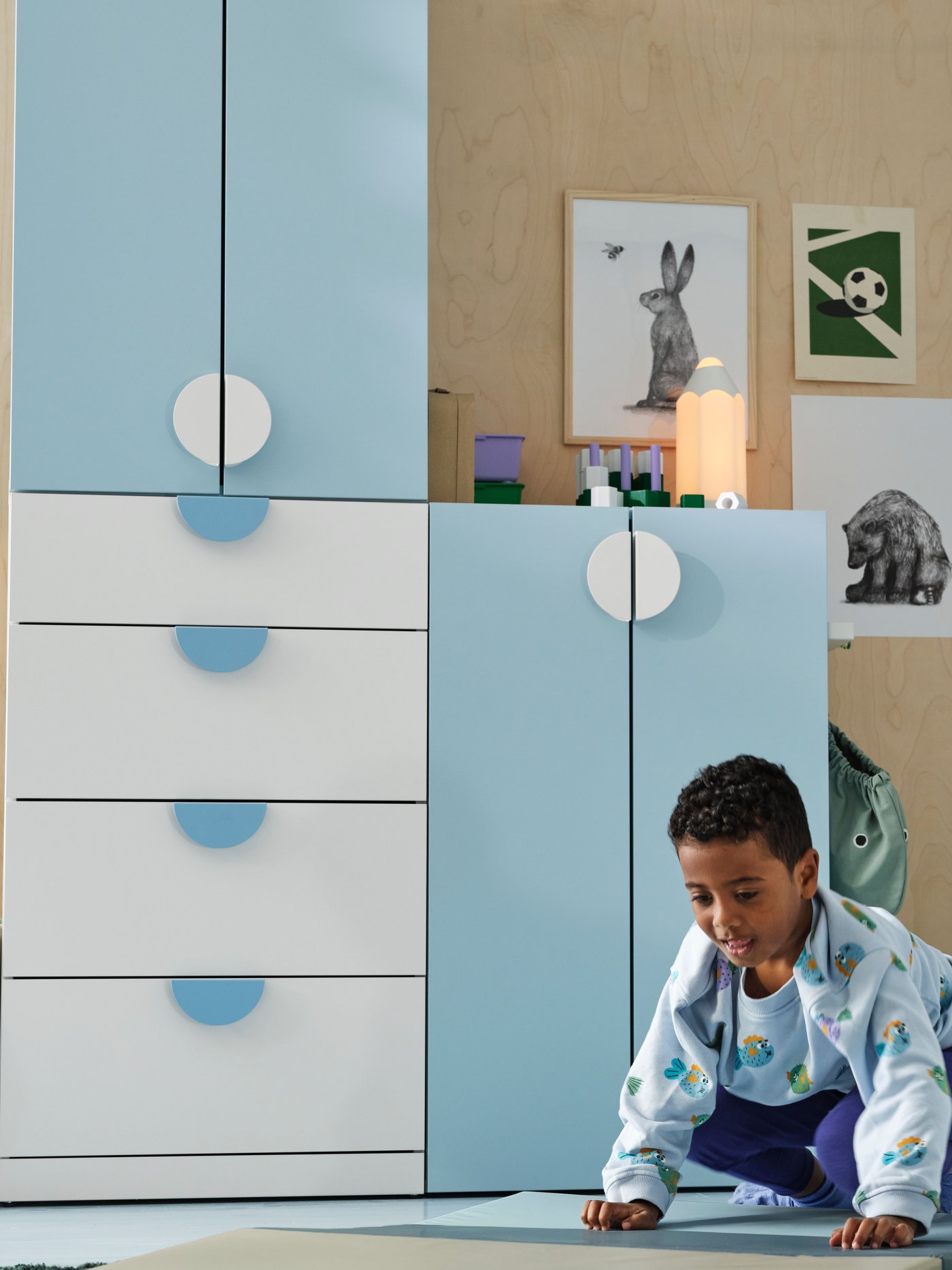 A child on a blue/green mat in a room with a light blue and white LASTARE storage combination, animal posters on the wall.