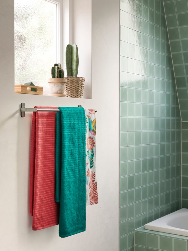 Green and pink towels with woven stripes hang on a BROGRUND towel rail that’s wall-mounted under a window.