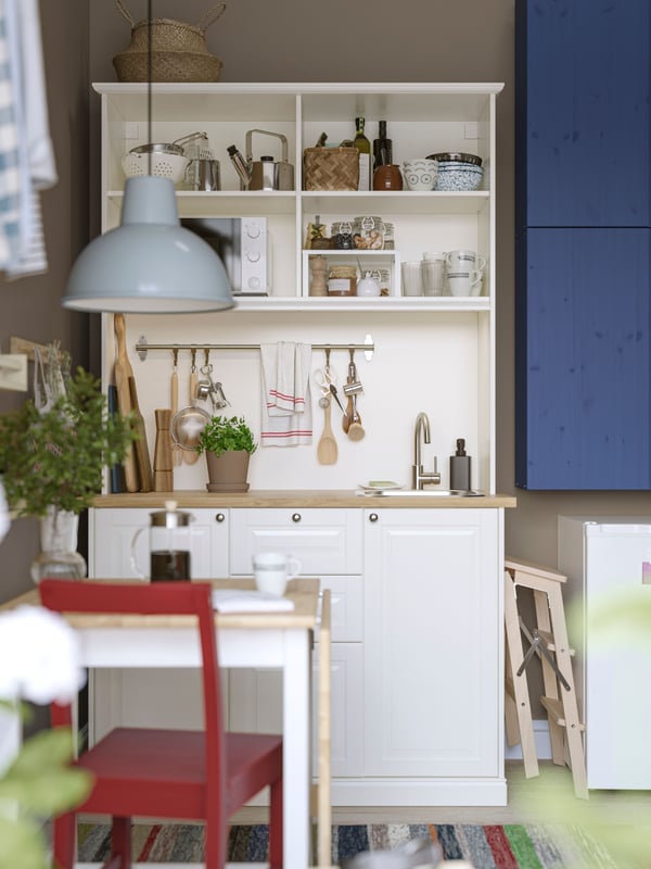 A white ÖNNERUP kitchenette has blue-painted IVAR cabinets mounted next to it and a small table with a red chair in front.