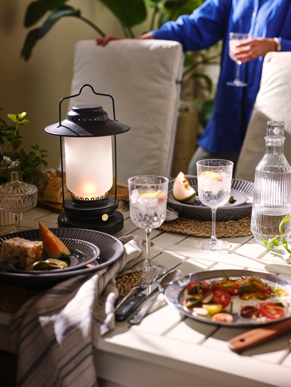 A close-up of a BONDHOLMEN outdoor table with some plates, glasses and a black STORHAGA LED table lamp on it.