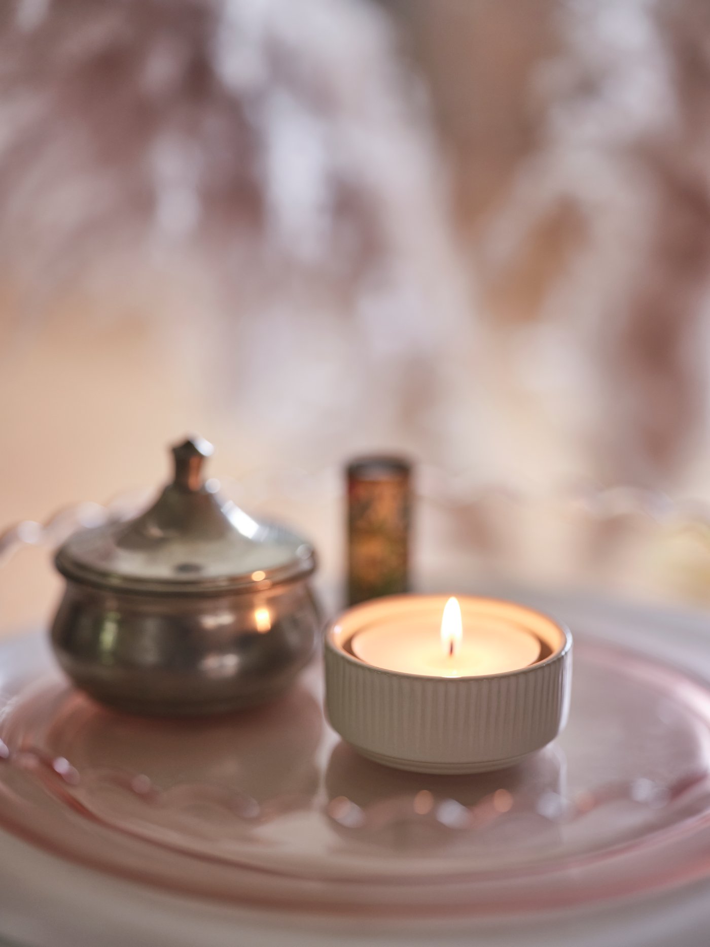 VÅRDANDE ceramic candle holder with lit candle and brass pot with lid sitting on a tabletop.
