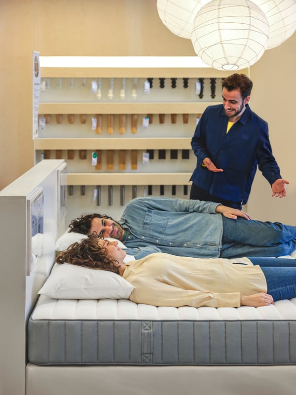 Two people lying side by side on a VALEVÅG mattress in an IKEA store. A store co-worker stands by the bed talking to them.