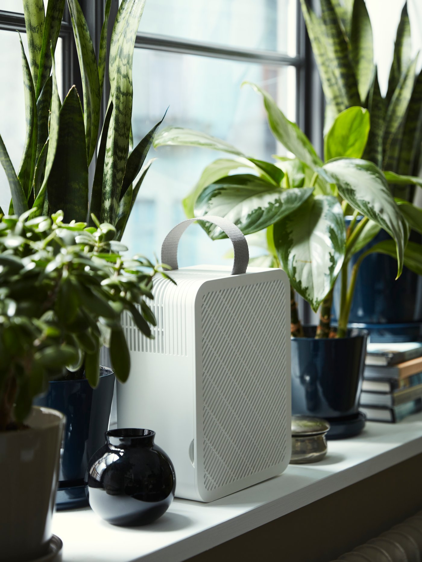 An UPPÅTVIND air purifier stands on a windowsill next to some plants in plant pots, books and other objects.