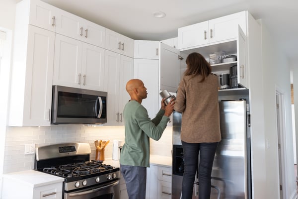 A couple are in their kitchen, tidying and placing things into an above fridge cupboard.