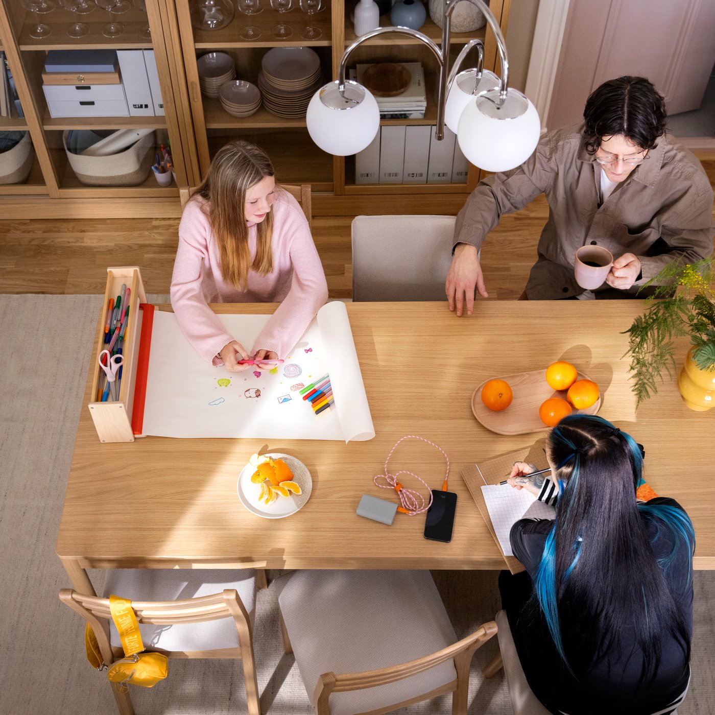 People sitting around a TONSTAD table in oak veneer, carrying gout activities like studying and drawing, fruit on the table.