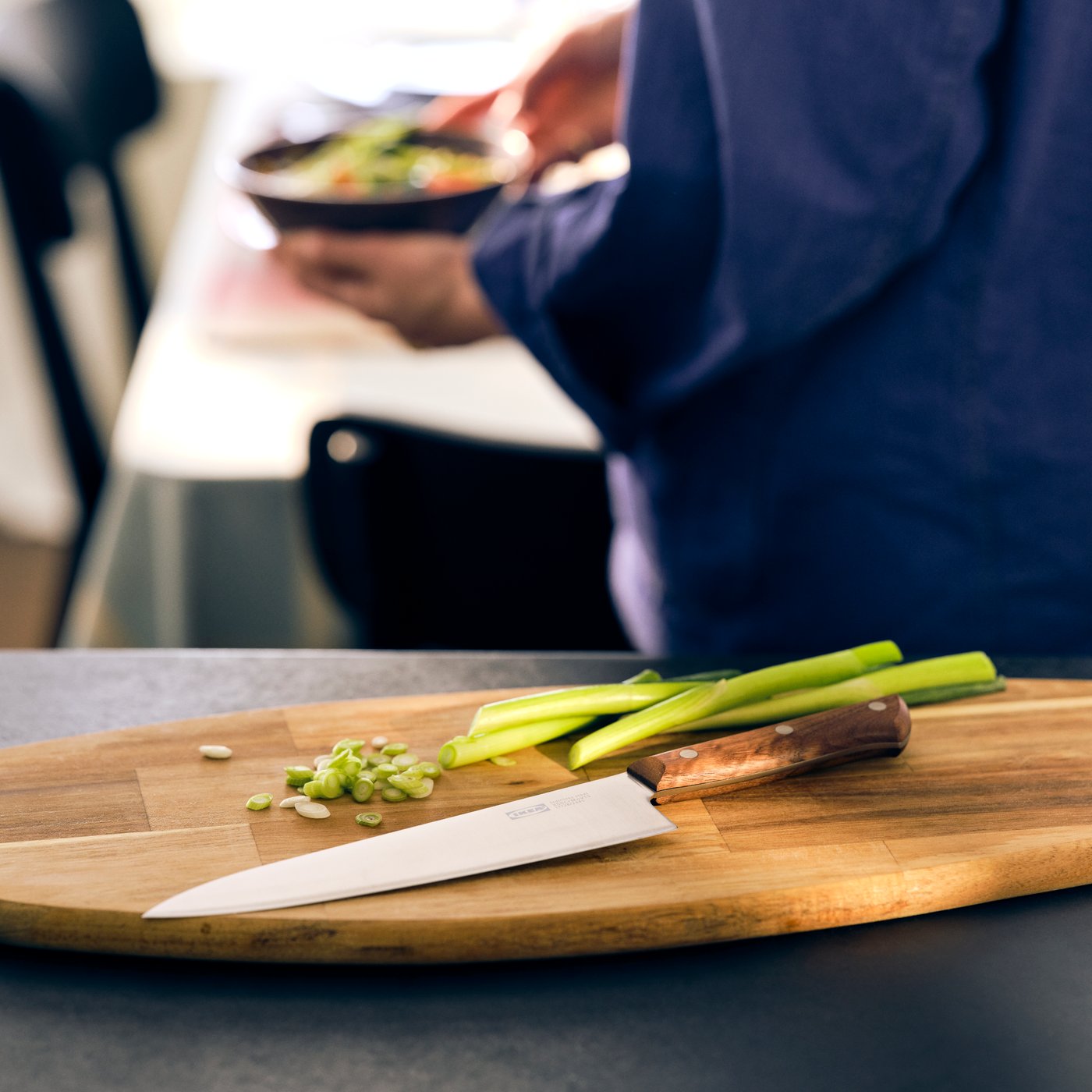 Couteau VARDAGEN avec manche en bois posé sur une planche à découper ovale, près de quelques oignons verts.
