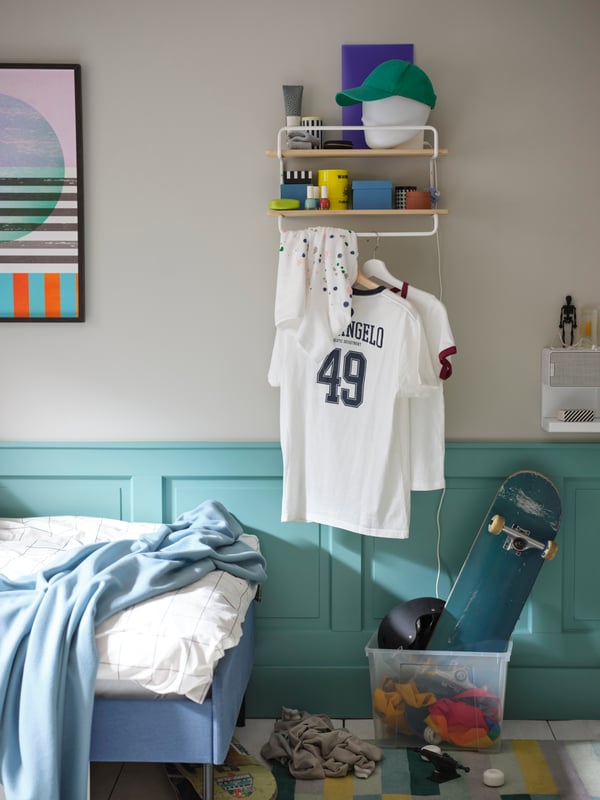 Une étagère murale SPORTSLIG en blanc/bouleau sur laquelle sont rangés différents objets et des vêtements sur des cintres; non loin, une boîte transparente contenant un skateboard.
