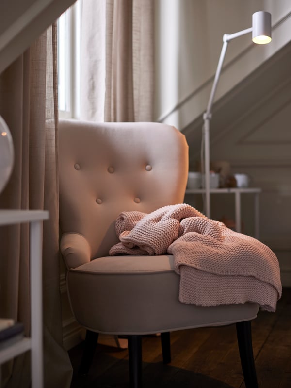 A pale pink knitted throw on a beige REMSTA armchair with a white floor lamp behind, by a window with beige curtains.