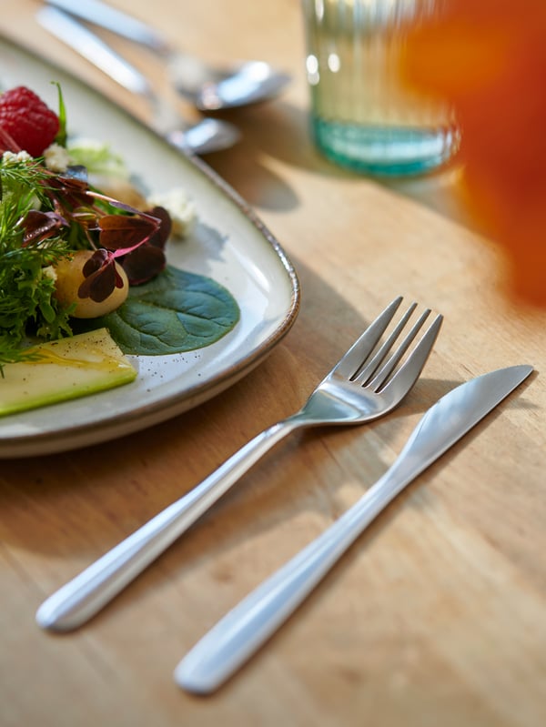 Un tenedor y un chuchillo FÖRNUFT junto a un plato GLADELIG con una ensalada servida, sobre una mesa de madera. 