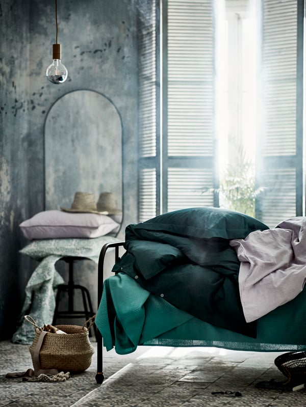 Une chambre à coucher lumineuse avec un lit orné de linge de lit vert. À côté se trouvent un miroir et un tabouret avec des oreillers supplémentaires.