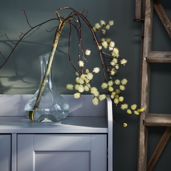 Une branchette sculpturale, solitaire et pleine de fleurs dans une carafe en verre transparent STORSINT, elle-même posée sur un buffet HAUGA gris.