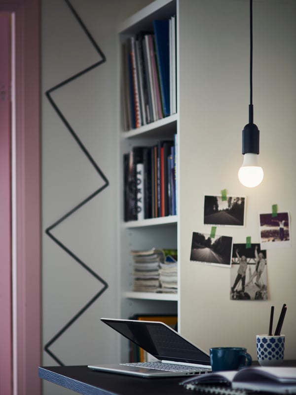 Lampadina a LED SOLHETTA accesa, appesa al soffitto sopra a un tavolo con PC portatile, una tazza e dei libri.