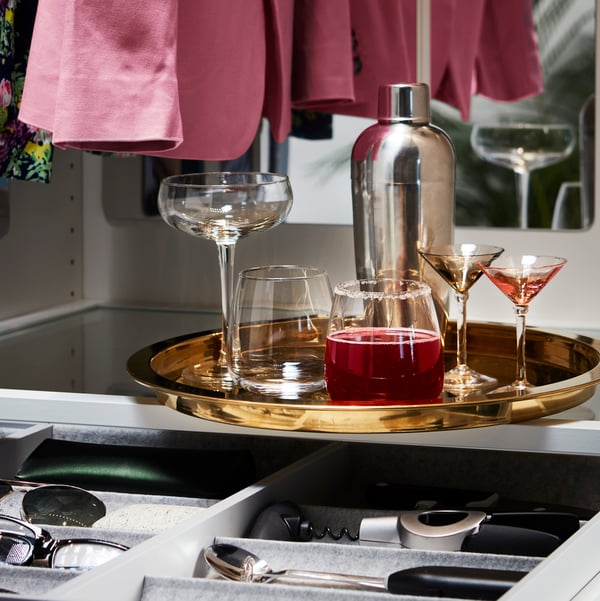 Bilden visar en bricka med olika glas och en cocktailshaker. Brickan är placerad ovanpå en låda som innehåller bestick och redskap. I bakgrunden syns några hängande kläder.
