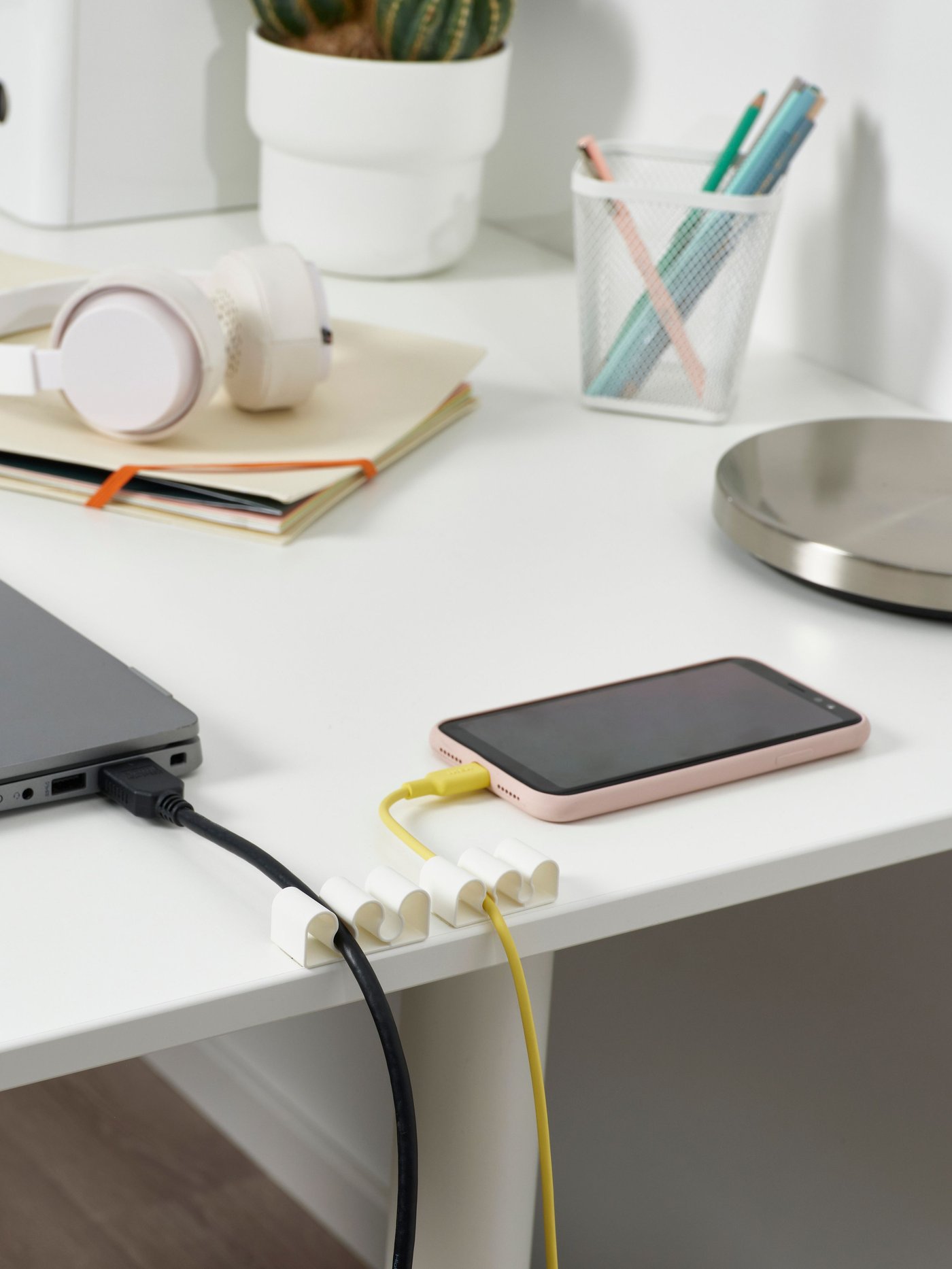 En bærbar computer og en smartphone ligger på et hvidt skrivebord med kabler tilsluttet en kabelholder.