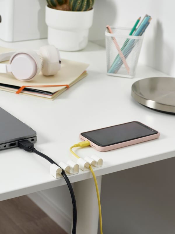 Two white KALKSTUFF cable holders are attached to a white desk, one with a laptop cable and one with a phone charging cable.