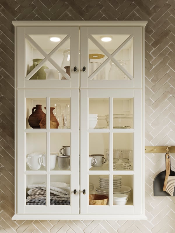 A cabinet with BODBYN glass doors in off-white holding diverse plates, cups, bowls, towels and other items on the shelving.