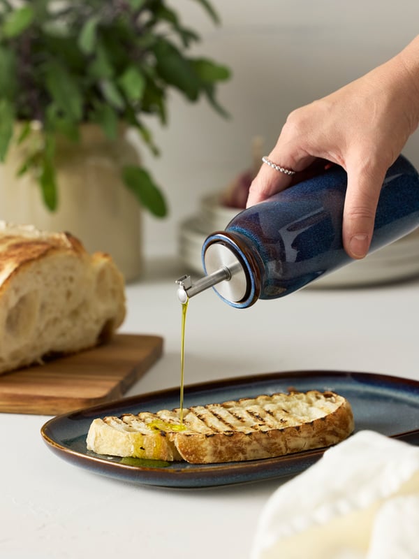 Рука держит синюю бутылку BRUGDHAJ, капая масло на кусок поджаренного хлеба на тарелке GLADELIG.