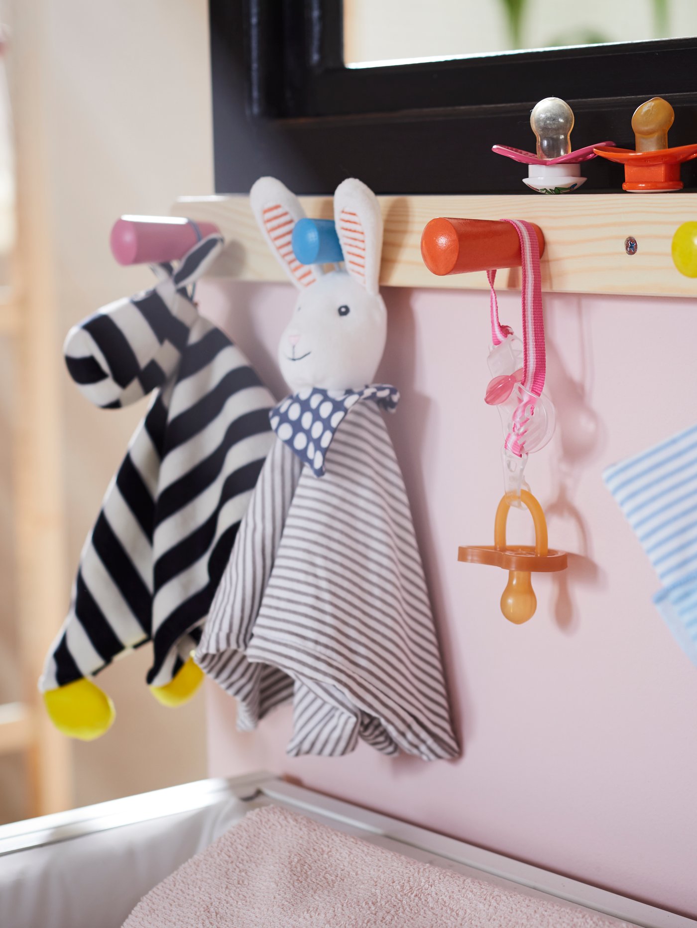 A FLISAT knob rack with 4 knobs is attached to a pink wall, holding KLAPPA comfort blankets with soft toys and soothers.