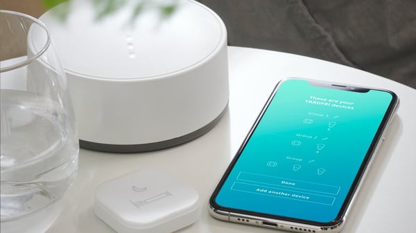TRÅDFRI gateway lying on a bedside table together with TRÅDFRI remote and the IKEA Home smart app displayed on a mobile device.