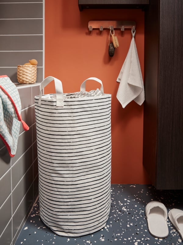 Una cesta de ropa sucia KLUNKA blanca con rayas negras, en la esquina de un baño con cerámicas grises y un muro naranjo. Junto a la bolsa hay unas pantuflas NJUTA y, un poco más arriba, cuelga desde una percha metálica una toalla pequeña VÅGSJÖN blanca.
