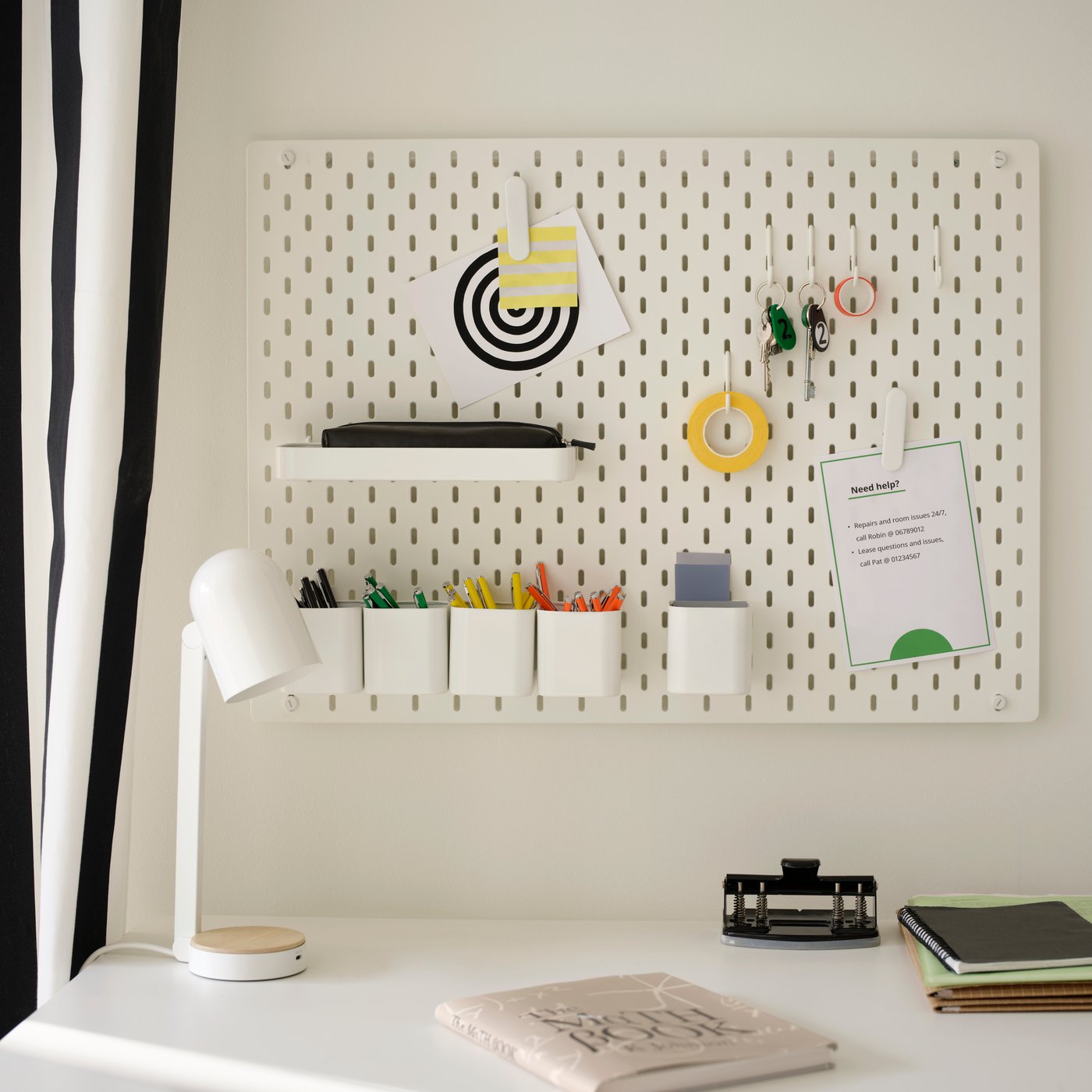 A white SKÅDIS pegboard combination is on a wall above a desk and has a note with tenant information in one of its clips.