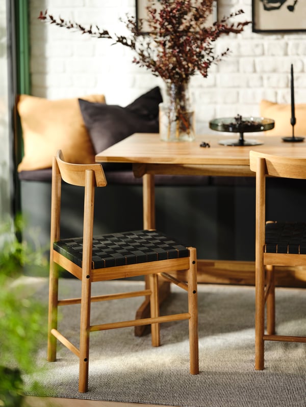A NACKANÄS acacia table, two NACKANÄS acacia chairs and a bench with cushions against a white brick wall.