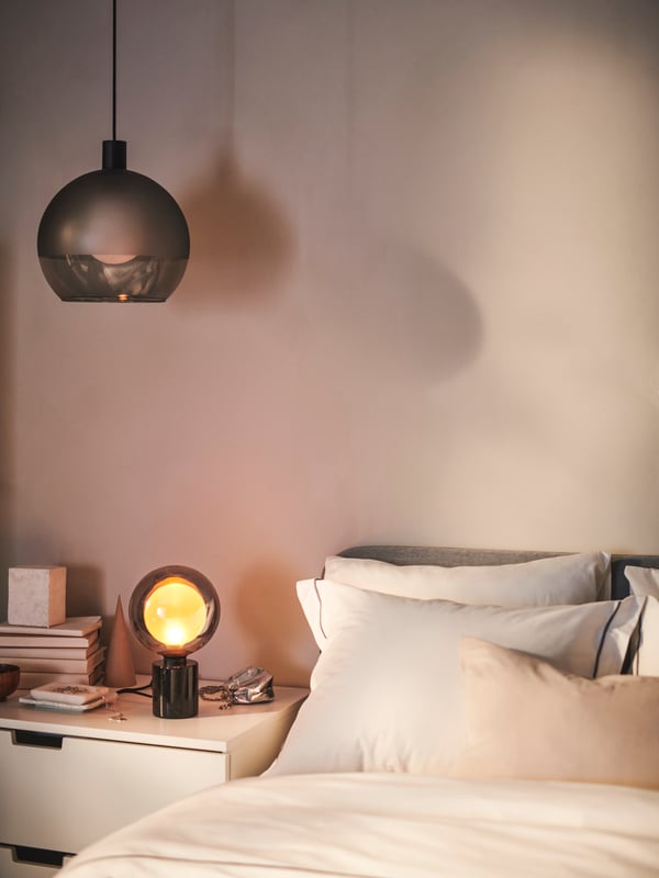 Eine JAKOBSBYN Hängeleuchte über einer Tischleuchte mit einem MOLNART LED-Leuchtmittel, die auf einer Kommode neben einem Bett steht.