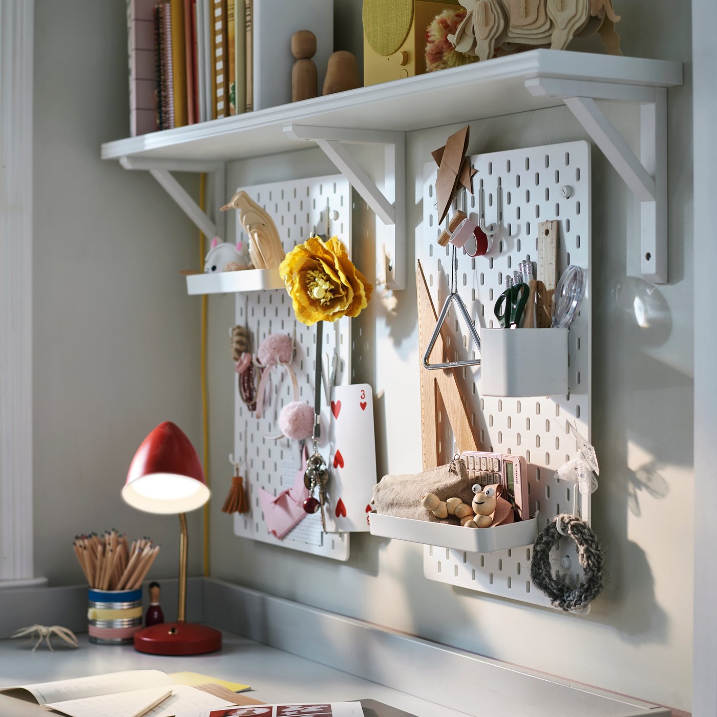 Two white SKÅDIS pegboard combinations on a wall hold stationery and accessories with a red ISNÅLEN LED work lamp nearby.