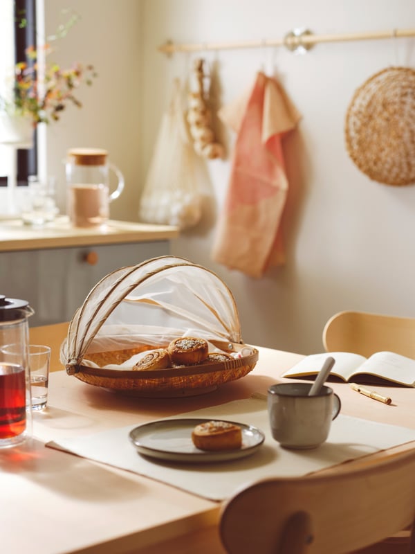 A table with a SNUSTORR food cover with tray in handmade bamboo holding KAFFEREP cinnamon buns by a plate with another bun.
