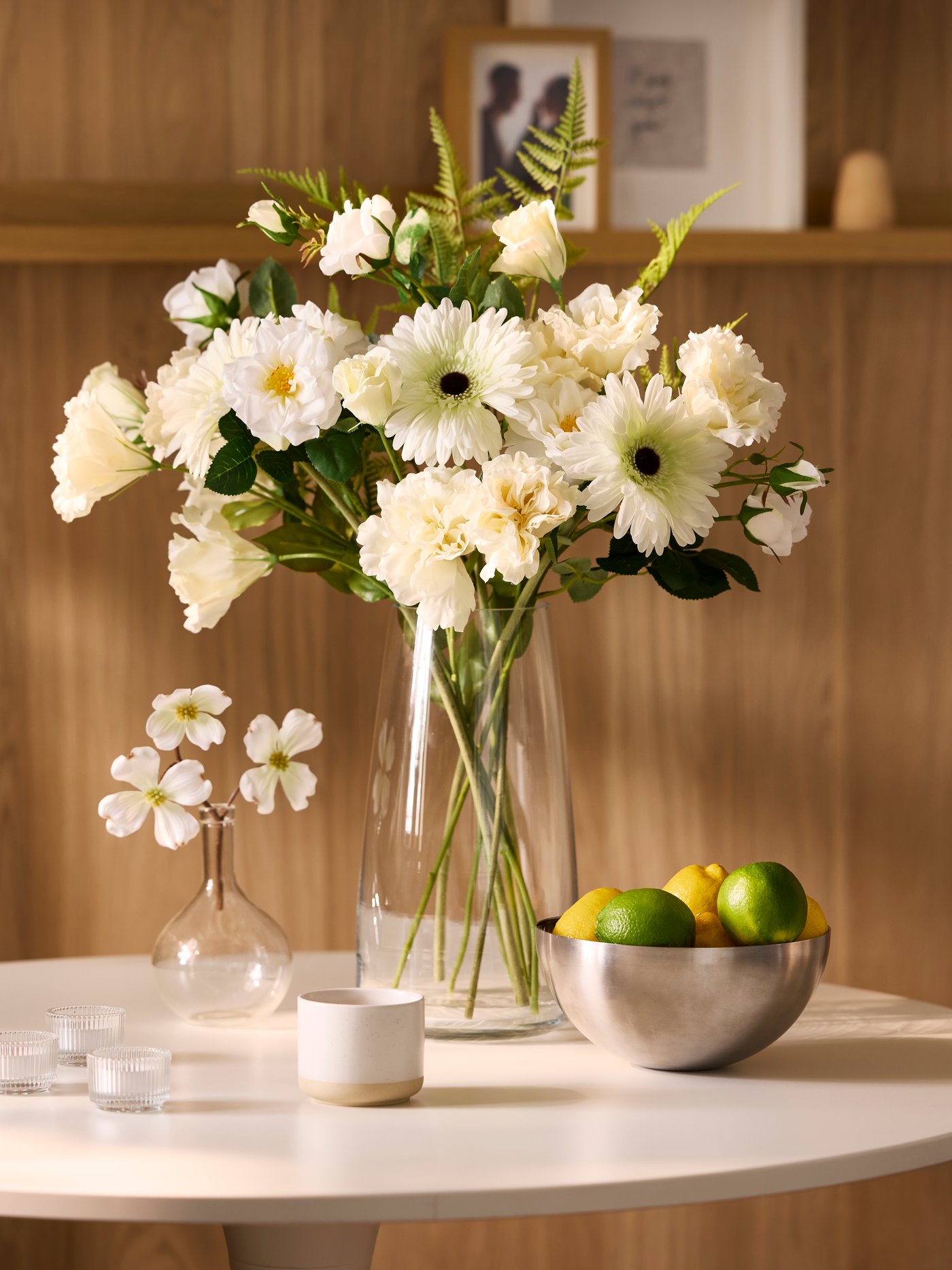 A sun-dappled dining area with a light-wood-panel wall and a white, round DOCKSTA table decorated with flowers and fruit.