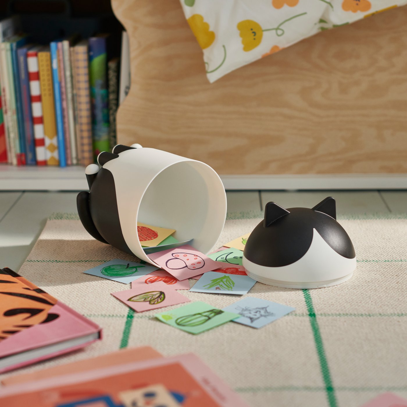 A cat-shaped GREJSIMOJS container lies opened on a carpet, revealing its content: a child’s paper cards.