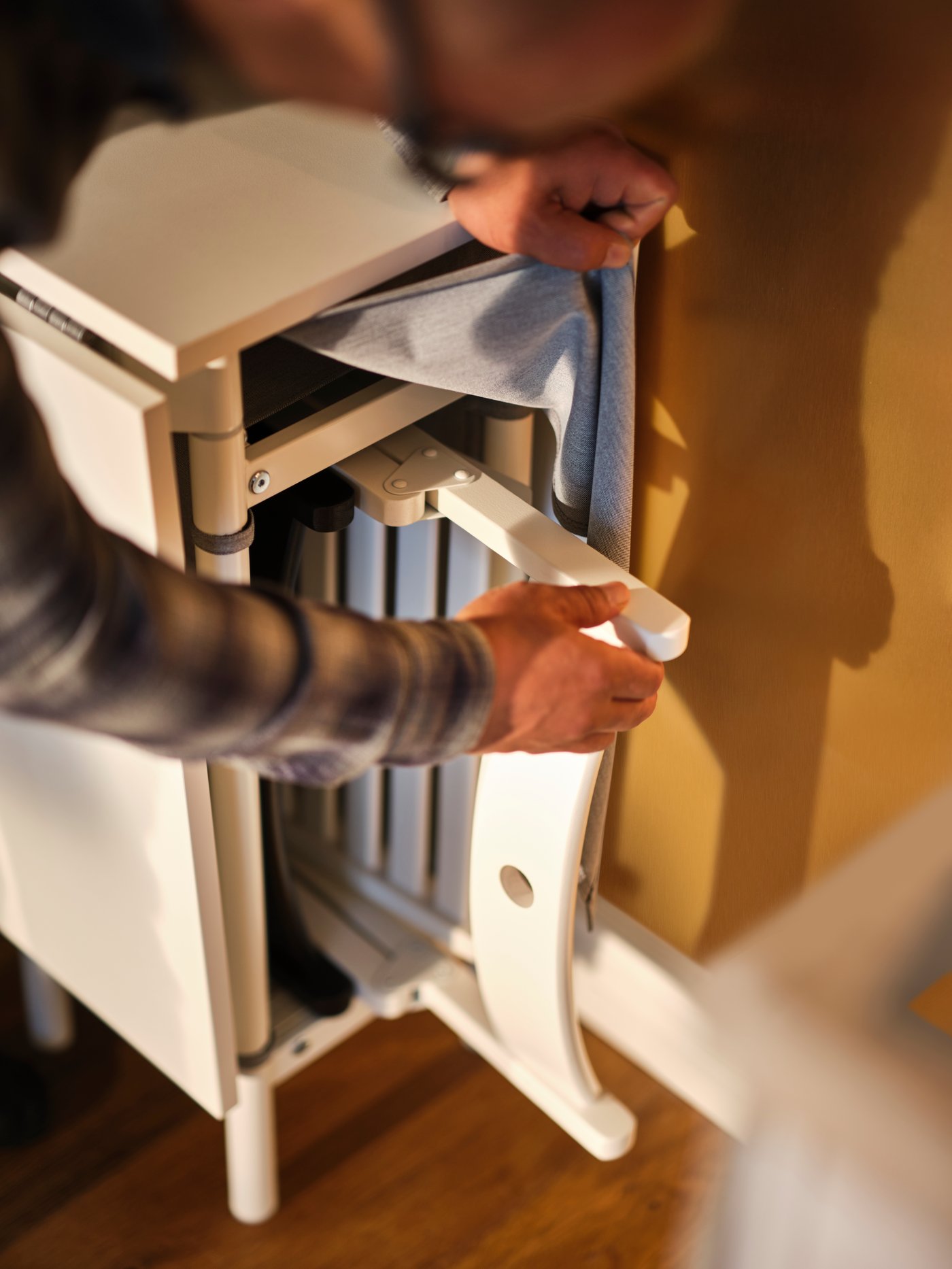 A person putting away a folded chair under a white/light grey KALLHÄLL gateleg table with storage against a wall.