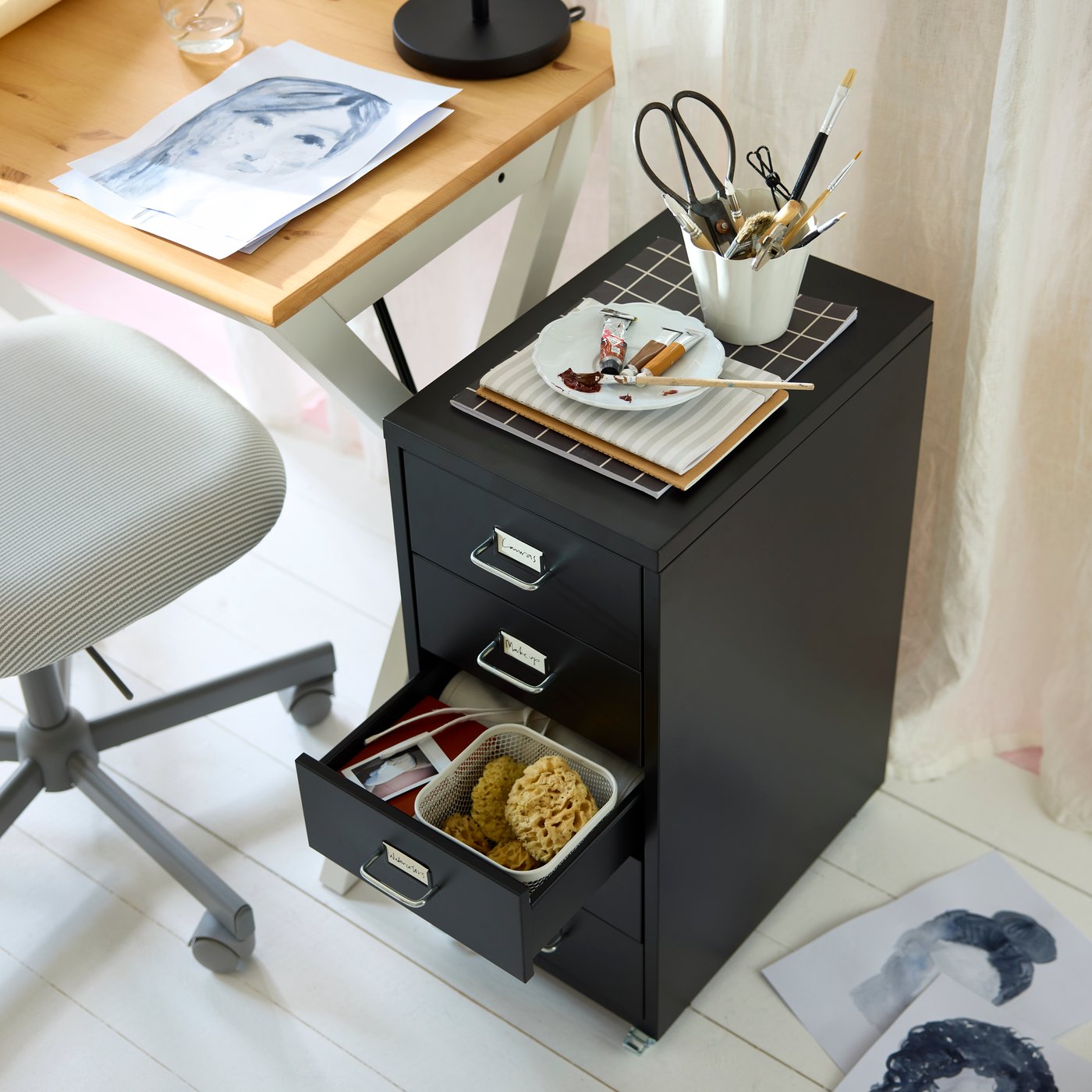 Black HELMER drawer unit on castors beside a BOLLPOJKE desk, holding art supplies with one drawer open.