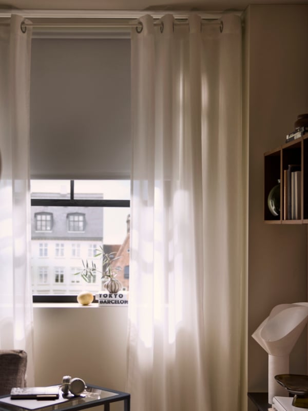 A window with a FRIDANS block-out roller blind and curtains partly shut, dimming the interior of the room inside.