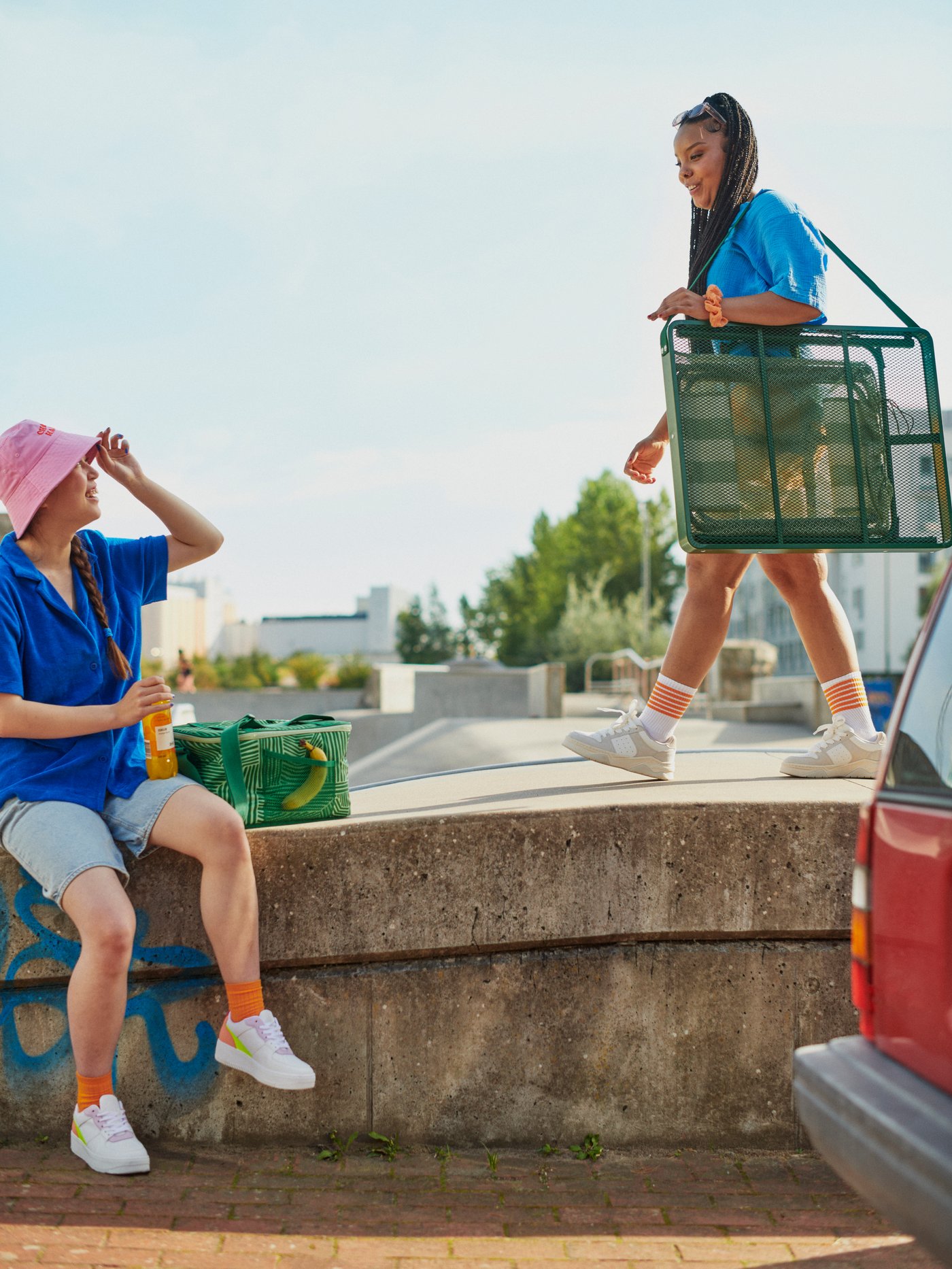 Young people in a sunny city landscape. One carries a STRANDÖN folding table set, one sits next to a NÄBBFISK cooling bag.