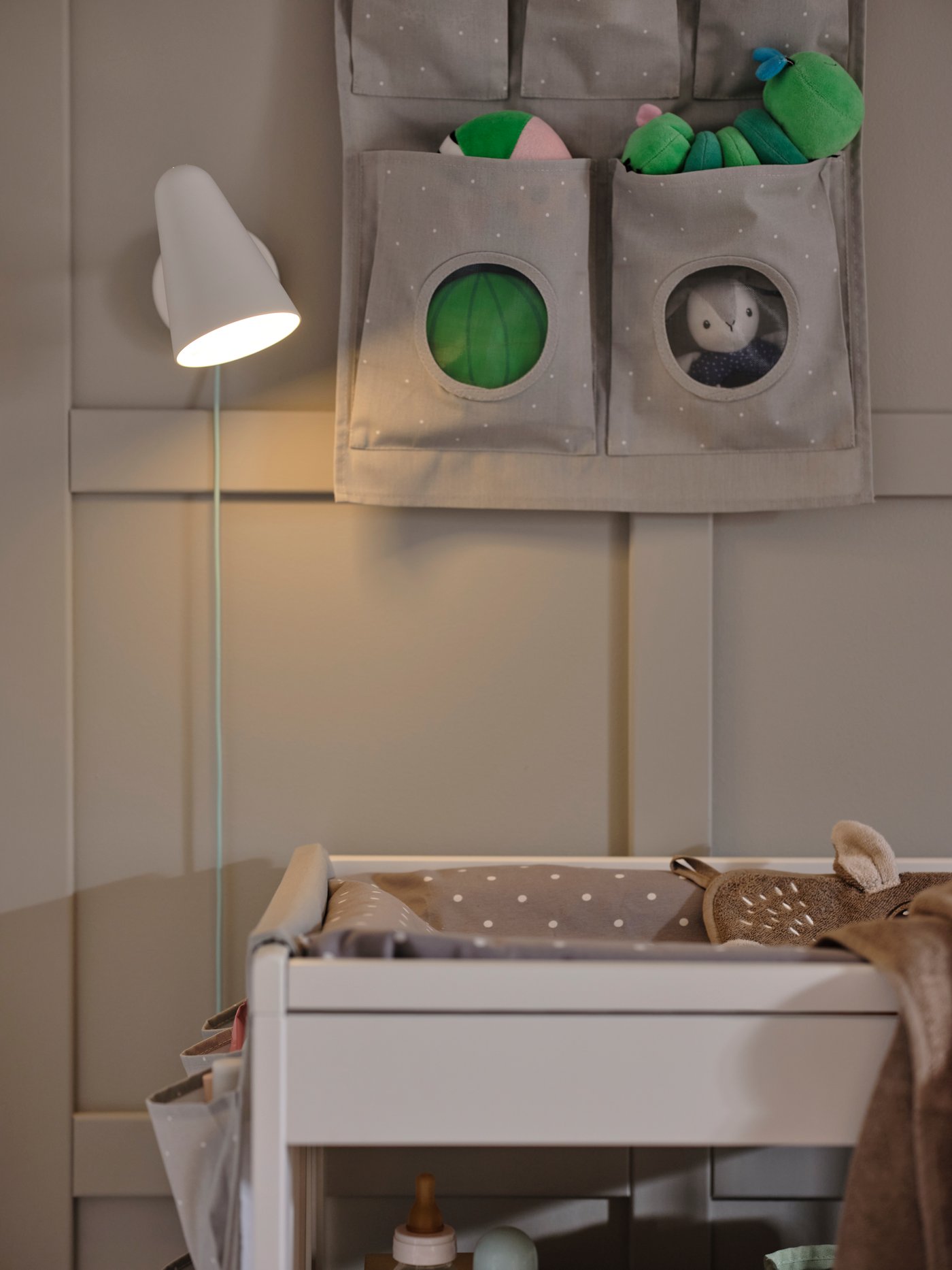 A white FUBBLA wall lamp with the bulb switched on above a GULLIVER changing table and with a LEN hanging storage beside it.