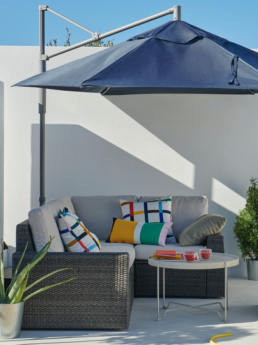 Un quitasol LINDÖJA cubre un living exterior con un sillon SOLLERÖN en L con cojines crema y rayados de colores, y una mesa de centro blanca KROKHOLMEN, frente a un muro blanco.